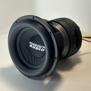 May include: A black Sundown Audio subwoofer with a carbon fiber cone. The subwoofer has a circular design with a prominent speaker cone and the brand name in white. The unit is designed for high-performance audio systems.