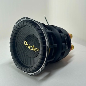 Op de afbeelding: Een zwarte subwoofer-luidspreker met een textuur van koolstofvezel op de conus, waarop het woord Pride in het geel is gedrukt. De luidspreker heeft een dikke, geribbelde rubberen rand, een stevig frame en goudkleurige aansluitingen aan de achterkant.