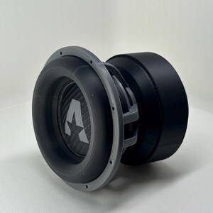 May include: A circular subwoofer speaker featuring a grey frame and a black cone with a carbon fiber texture. The center of the cone displays a prominent white letter A logo. The back of the unit consists of a large, solid black cylindrical magnet housing.