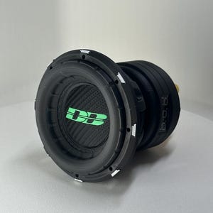 Op de afbeelding: Een zwarte subwoofer met een koolstofvezel conus en een groen "DB" logo. De subwoofer heeft een cirkelvormig ontwerp met een reeks witte accenten rond de rand. De zijkant van de subwoofer heeft de tekst "B.O.B."
