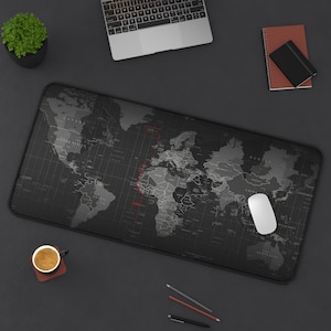 May include: A large, rectangular black and gray world map mouse pad with a white computer mouse. The map shows country outlines and grid lines. A laptop, coffee cup, and small plant are also on the dark surface.