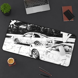 May include: Black and white desk mat with a manga-style illustration of two sports cars racing. The design includes a driver in one car. A computer mouse sits on the mat, alongside a laptop and other desk accessories.
