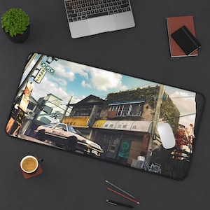 Op de afbeelding: Zwarte rechthoekige bureaumat met een gedetailleerde illustratie van een Japanse straatscène. De afbeelding toont een witte auto, gebouwen, straatnaamborden en een blauwe lucht. Een laptop, muis en koffiekopje zijn ook zichtbaar.
