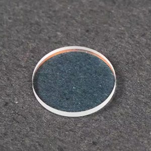 Puede incluir: Una peque&ntilde;a lente de vidrio redonda y transparente con un borde blanco y un anillo interior naranja. El centro de la lente tiene una apariencia azul oscuro texturizada. La lente est&aacute; sobre un fondo gris oscuro.