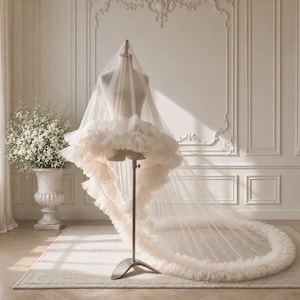 May include: A long, ivory-colored tulle wedding veil draped over a mannequin. The veil features a voluminous, multi-layered ruffled trim along the entire edge, creating a circular, flowing silhouette that pools onto the floor in a soft, sheer fabric.