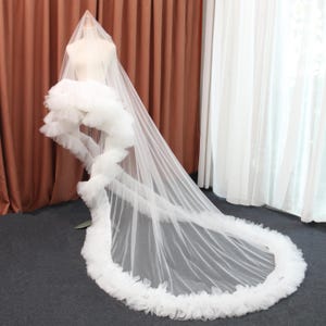 May include: A white bridal veil with a long train, displayed on a mannequin. The veil features a cascading ruffle design along the train's edge and a layered bodice. The sheer fabric creates a flowing, elegant look for a wedding.