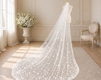 Puede incluir: Un velo de novia blanco largo hasta el suelo hecho de tela de tul transparente, con aplicaciones florales blancas tridimensionales dispersas que se vuelven más densas hacia el dobladillo redondeado, exhibido en un torso de maniquí.