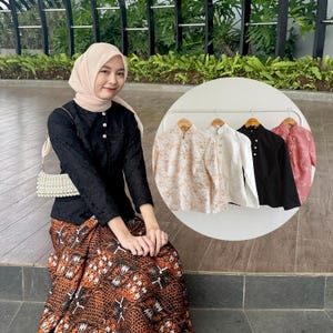 May include: A long-sleeved black blouse with a textured floral pattern, a high collar, and white buttons, paired with a brown and black patterned batik skirt. A circular inset shows four similar blouses in beige, white, black, and pink on hangers.