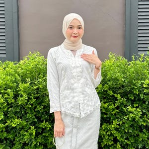 Peut inclure: Ensemble deux pièces blanc brodé, composé d'un haut à manches longues et d'une jupe. Le haut présente des motifs floraux complexes et une fermeture boutonnée. La jupe a un design assorti. L'ensemble est porté avec un voile assorti.