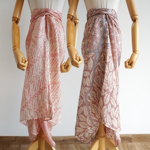 Könnte beinhalten: Zwei Sarongs, die auf Holzschaufensterpuppen drapiert sind. Der linke Sarong zeigt ein hellrosa-weißes Batikmuster mit sich wiederholenden geometrischen Formen. Der rechte Sarong zeigt ein malvenfarbenes Blattmotiv mit einem detaillierten Druck.