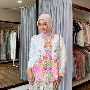 Puede incluir: Una blusa blanca de encaje de manga larga con bordados florales vibrantes en tonos rosa, naranja y verde a lo largo de la parte delantera. La prenda se combina con un pañuelo rosa claro y una falda estampada con motivos verdes y rosas.