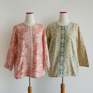 May include: Two long-sleeved shirts displayed on mannequins. The left shirt is coral with a white floral pattern and a central vertical geometric stripe. The right shirt is beige with a teal floral pattern and a central vertical geometric stripe.