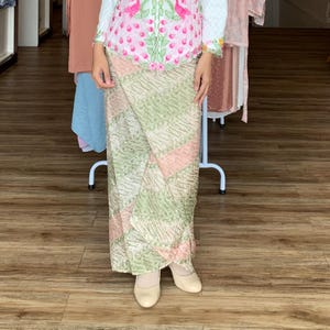 May include: A long wrap skirt featuring a diagonal batik pattern in shades of sage green and soft pink. The skirt is paired with a white long-sleeved top decorated with embroidered pink flamingos and green foliage along the front centre panel.