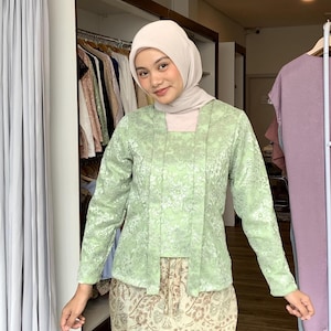 May include: A light green long-sleeved top featuring a floral lace overlay with a square neckline and two vertical front panels. The garment is paired with a beige headscarf and a patterned skirt with brown and cream botanical motifs.