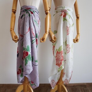 May include: Two floral sarongs, one in lavender and the other in white, displayed on wooden mannequins. The lavender sarong has red and green flowers, and the white one features red and green floral patterns. Each is wrapped around the mannequin's waist.