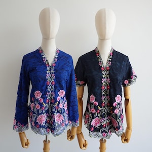 May include: Two lace blouses featuring V-necklines and floral embroidery in shades of pink, green, and white. The left blouse is royal blue with long sleeves, while the right blouse is black with short sleeves. Both garments have scalloped hems.