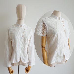 May include: A white long-sleeved blouse featuring a delicate diamond-patterned lace overlay, a mandarin collar, and an asymmetrical front closure secured with three small round pearl-like buttons, displayed on a mannequin.