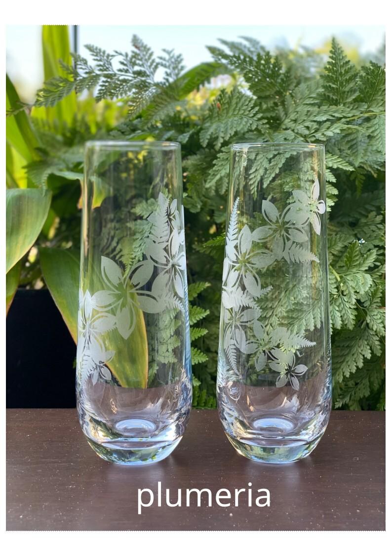 May include: Two clear glass tumblers with etched white plumeria flowers. The glasses are on a wooden surface.