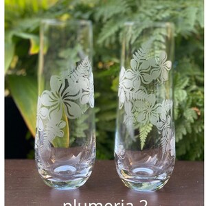 May include: Two clear glass champagne flutes with etched white plumeria flower designs. The glasses are on a dark brown wooden surface. The text "plumeria 2" is printed below the glasses.