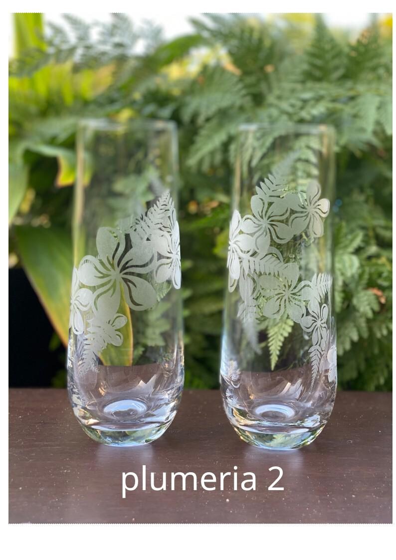 May include: Two clear glass champagne flutes with etched white plumeria flower designs. The glasses are on a dark brown wooden surface. The text "plumeria 2" is printed below the glasses.