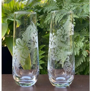 May include: Two clear glass tumblers with etched hibiscus flower designs. The glasses are on a dark brown wooden surface.