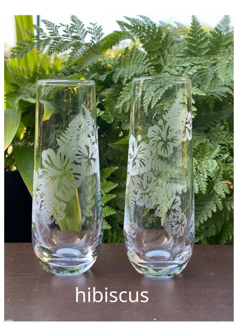 May include: Two clear glass tumblers with etched hibiscus flower designs. The glasses are on a dark brown wooden surface.