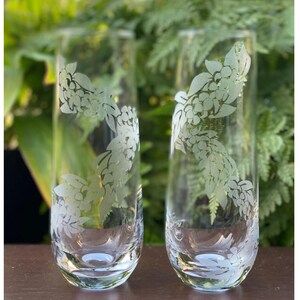 May include: Two clear glass tumblers with a floral design etched into the glass. The design is of white flowers and leaves. The glasses are sitting on a wooden surface.