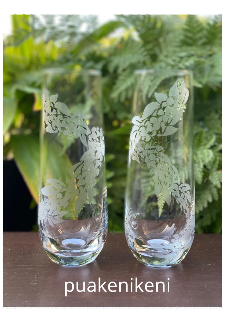 May include: Two clear glass tumblers with a floral design etched into the glass. The design is of white flowers and leaves. The glasses are sitting on a wooden surface.