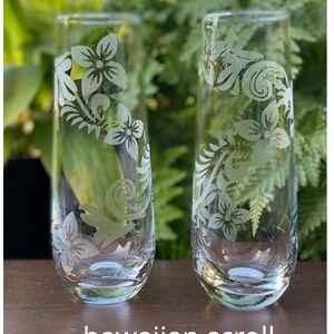 May include: Two clear glass tumblers with etched floral designs. The design features a scrolling pattern of flowers and leaves. The glasses are on a wooden surface. The text "hawaiian scroll" is below the glasses.