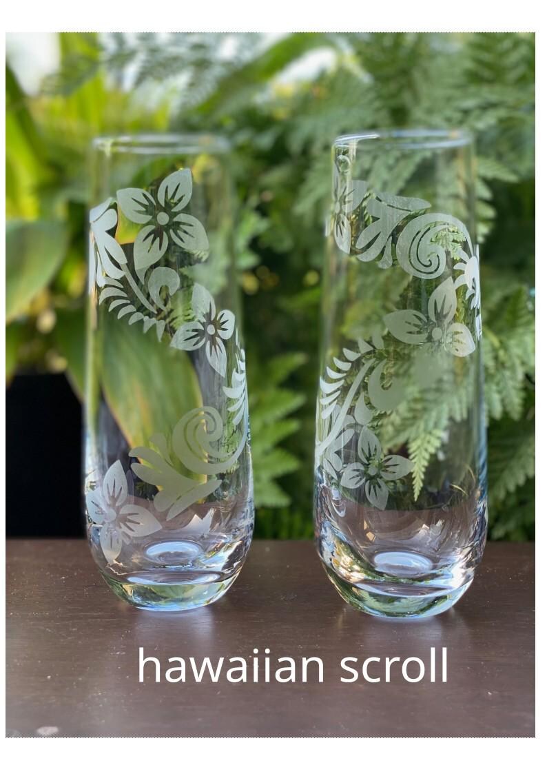 May include: Two clear glass tumblers with etched floral designs. The design features a scrolling pattern of flowers and leaves. The glasses are on a wooden surface. The text "hawaiian scroll" is below the glasses.