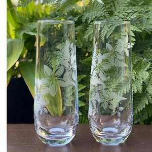 May include: Two clear glass champagne flutes with etched floral designs. The flutes are on a wooden surface.