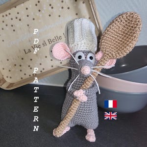 以下が含まれることがあります： 白いコック帽をかぶり、ベージュの木のスプーンを持った、かぎ針編みの灰色のネズミ。 ネズミはピンクの耳と足、白いひげがあります。 画像には「PDF Pattern」というテキストが含まれています。