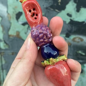 May include: A colourful ceramic fruit-shaped object with a red strawberry base, a blue berry, a purple cluster, and a red top with holes. The object has gold and green accents and is held in a hand.