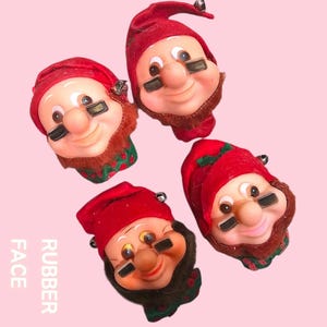 May include: Four vintage rubber face Christmas elf ornaments. Each elf has a red hat with a bell, a painted face with black eye paint, and a brown beard. The text "RUBBER FACE" is printed vertically on the left side.