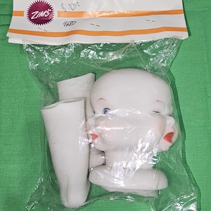 May include: A vintage doll head and arm parts are packaged in a clear plastic bag. The doll head is white with blue eyes and red lips. The packaging includes the ZIMS logo and handwritten text.
