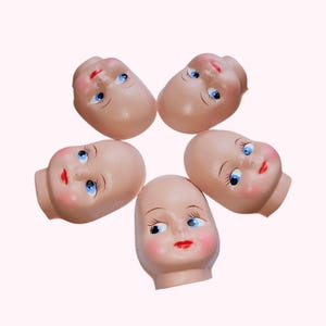 May include: Five vintage doll heads arranged in a circular pattern. The doll heads are made of a flesh-toned material, with blue eyes, red lips, and rosy cheeks. Each head has painted eyelashes and eyebrows.