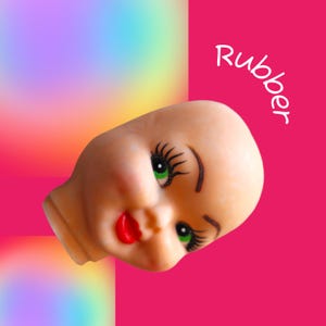May include: A rubber doll head with green eyes, long black eyelashes, and red lips. The doll head is set against a vibrant pink background with a rainbow gradient in the upper left corner. The word "Rubber" is written in white.