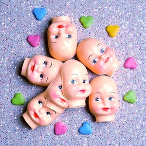 May include: A collection of vintage doll heads with painted features, including red lips and blue eyes, arranged with colorful heart and clover-shaped embellishments on a glittery surface. The doll heads are a pale peach color.