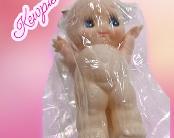 Könnte beinhalten: Eine Vintage-Kewpie-Puppe, verpackt in klarem Kunststoff. Die Puppe hat einen pfirsichfarbenen Hautton, blaue Augen und einen kleinen, lächelnden Mund. Die Puppe steht mit ausgestreckten Armen. Das Wort "Kewpie" ist in rosa Schreibschrift oben links geschrieben.