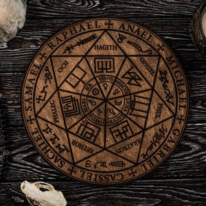 Puede incluir: Una pieza decorativa circular de madera con símbolos y texto grabados, incluyendo nombres como Rafael y Miguel. La imagen también presenta velas encendidas, un rosario, una calavera pequeña y un cráneo de ave, todo dispuesto sobre una superficie oscura con vetas de madera.