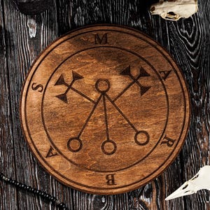 May include: A wooden, circular sigil with engraved symbols and the letters M, A, S, R, and B. The sigil is surrounded by candles, crystals, a bird skull, and a beaded necklace. The items are arranged on a dark wooden surface.