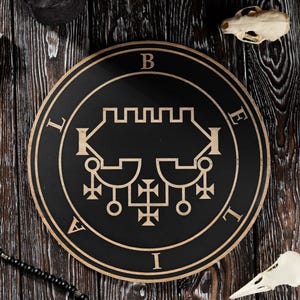 May include: A black and gold circular sigil with complex symbols, encircled by the letters B, E, L, I, and A. The image features a purple crystal, a bird skull, a candle, and a beaded necklace, set on a dark wooden surface.