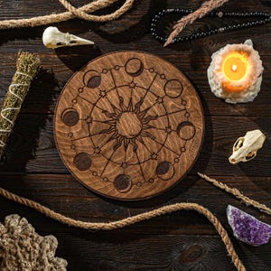 Könnte beinhalten: Ein rundes Holztablett mit einem Sonnen- und Monddesign. Das Tablett hat eine dunkelbraune Holzoptik und zeigt eine Sonne mit Strahlen und einen Kreis aus Monden in verschiedenen Phasen. Das Tablett ist von verschiedenen Gegenständen umgeben, darunter eine Kerze, ein Seil und ein Kristall.