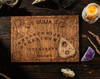 May include: A wooden Ouija board with a planchette in the shape of a heart. The board features the letters of the alphabet, numbers 1-9, and the words "yes", "no", and "good bye" printed on it. The board is surrounded by various objects, including a skull, a candle, and a crystal.