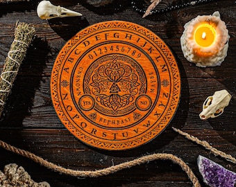 May include: A circular wooden Ouija board with engraved letters, numbers, and symbols. The board is orange with a tree of life design in the centre. A lit candle, crystals, and other mystical items surround the board.