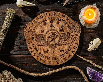 May include: A wooden Ouija board with engraved letters, numbers, and symbols, including an eye with wings. The board is surrounded by a lit candle, crystals, a bundle of herbs, and animal skulls, all arranged on a dark wooden surface.