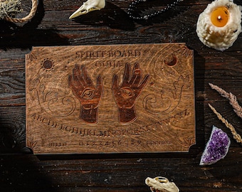 May include: A wooden spirit board with the words "Spirit Board Ouija" and the alphabet. The board features two hands with an eye design. Additional items include a candle, a skull, a crystal, and a beaded necklace.