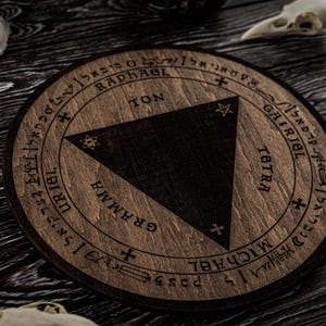 Op de afbeelding: Een ronde houten onderzetter met een donkerbruine driehoek in het midden. De onderzetter heeft gegraveerde tekst en symbolen langs de rand, waaronder de namen Raphael, Gabriel en Michael. Op een donker houten oppervlak.