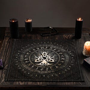 May include: A black and gold fabric altar cloth with a tree of life design and runes around the edge. The cloth is on a wooden table with black candles and a crystal.