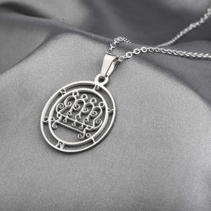 以下が含まれることがあります： 銀色のペンダントネックレス。楕円形のフレームの中に、複雑で様式化されたデザインが施されています。ペンダントは繊細なチェーンから吊り下げられており、グレーのテクスチャード加工された背景に配置されています。デザインには、装飾的で渦巻くパターンとシンボルが含まれています。